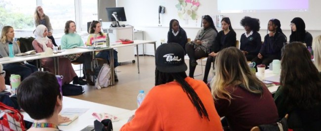 A group of young people sat in a row at the from of a classroom. Around them, sat at desks, are PGCE students, listening to the young people share their experiences of race, diversity and multiculturalism in school.